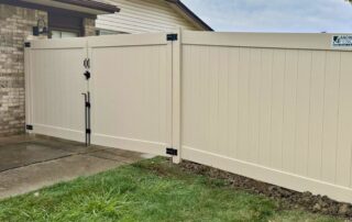 Cream colored vinyl privacy fence installed beside a brick house with a double gate featuring black hinges and latch hardware, enclosing a driveway on a residential property with grass and concrete visible in the foreground.