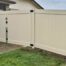 Cream colored vinyl privacy fence installed beside a brick house with a double gate featuring black hinges and latch hardware, enclosing a driveway on a residential property with grass and concrete visible in the foreground.
