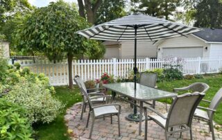 Backyard patio with a glass-top table and four chairs under a striped umbrella, surrounded by greenery, flowers, and a white picket fence with a residential home in the background.