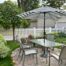 Backyard patio with a glass-top table and four chairs under a striped umbrella, surrounded by greenery, flowers, and a white picket fence with a residential home in the background.