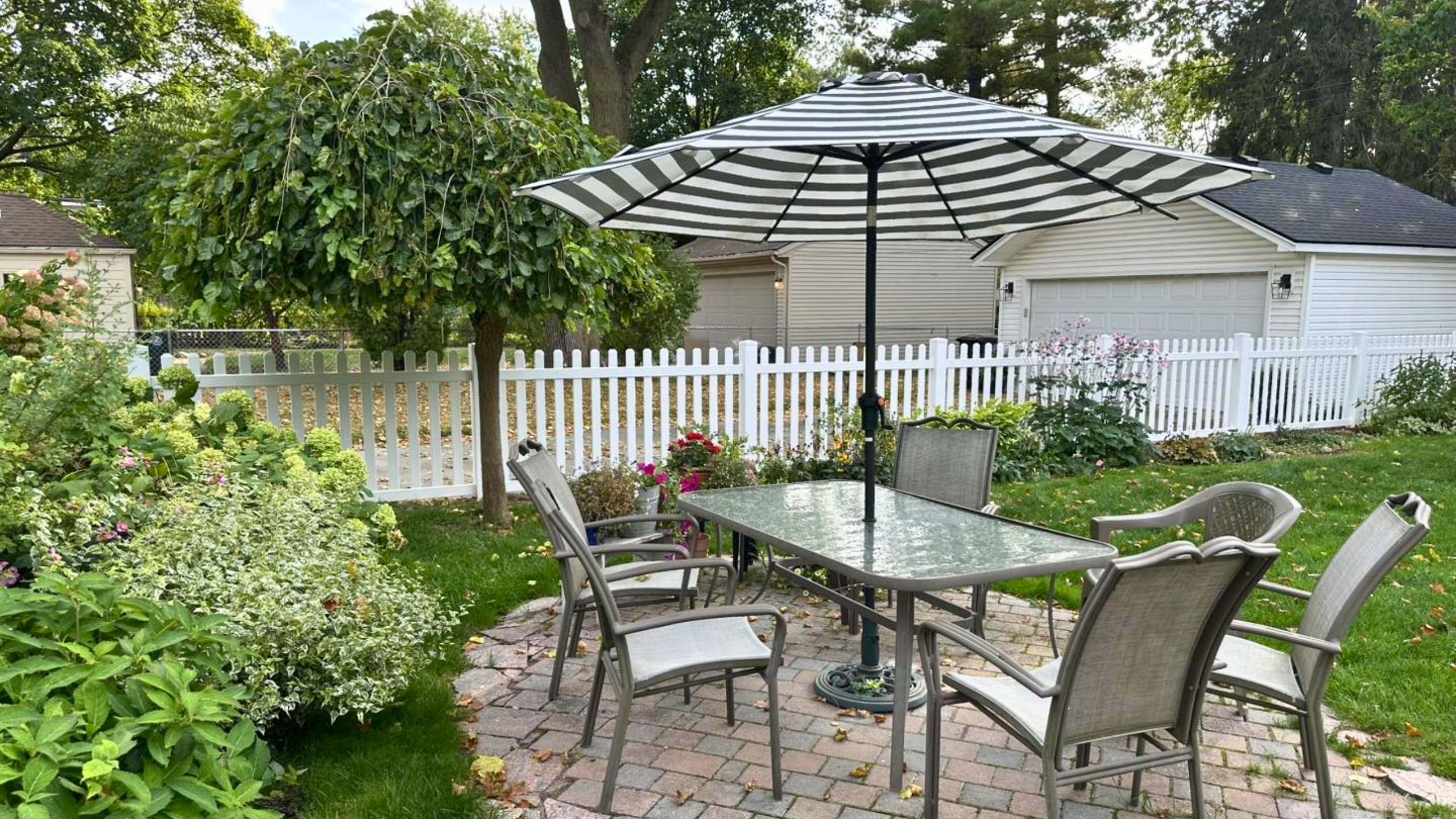 Backyard patio with a glass-top table and four chairs under a striped umbrella, surrounded by greenery, flowers, and a white picket fence with a residential home in the background.